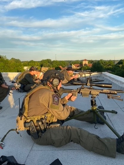 Sniper perched on a roof aiming down out of frame. Trees in background.